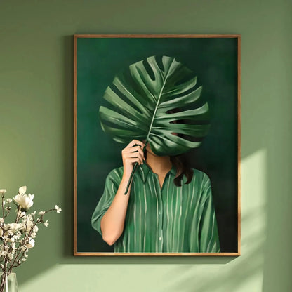 Framed artwork of a person holding a large green leaf over their face against a green wall.