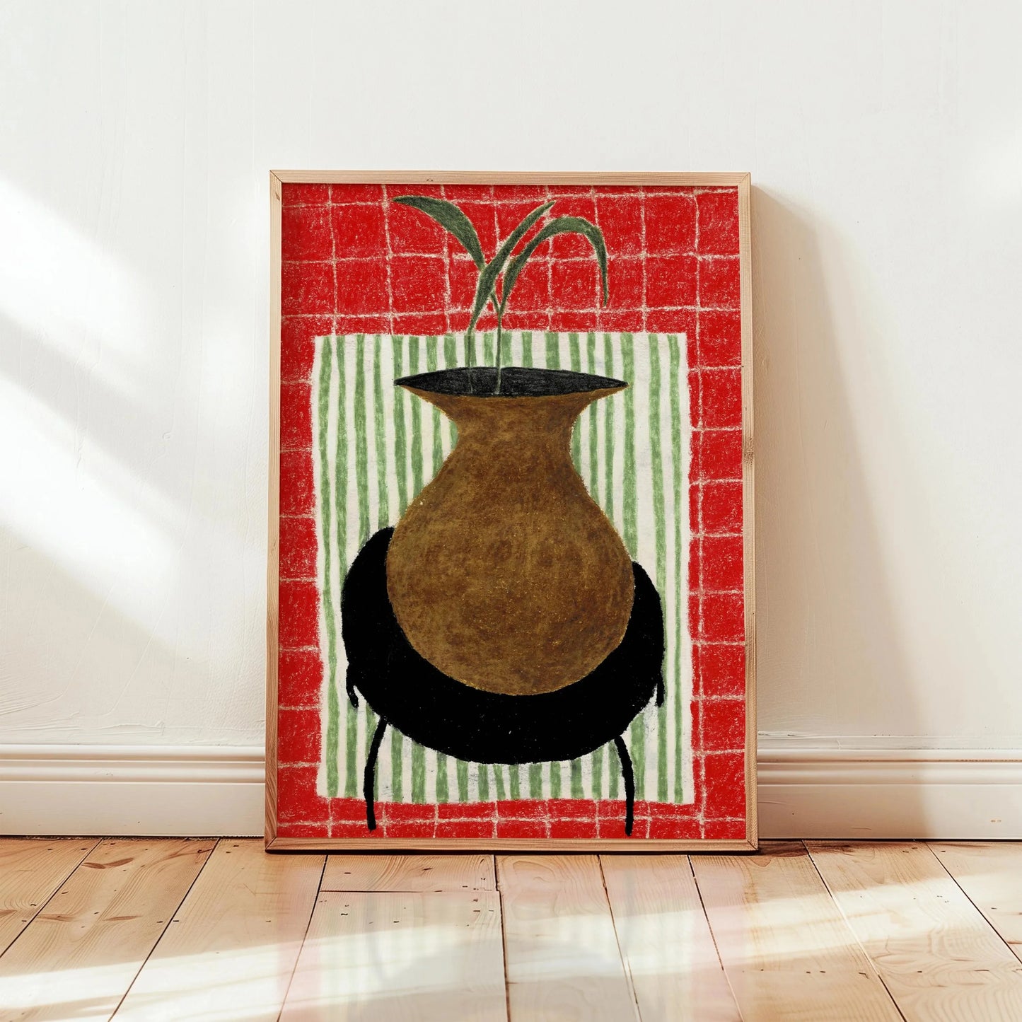 Framed artwork of a vase with a plant on a red and green striped background, leaning against a white wall.