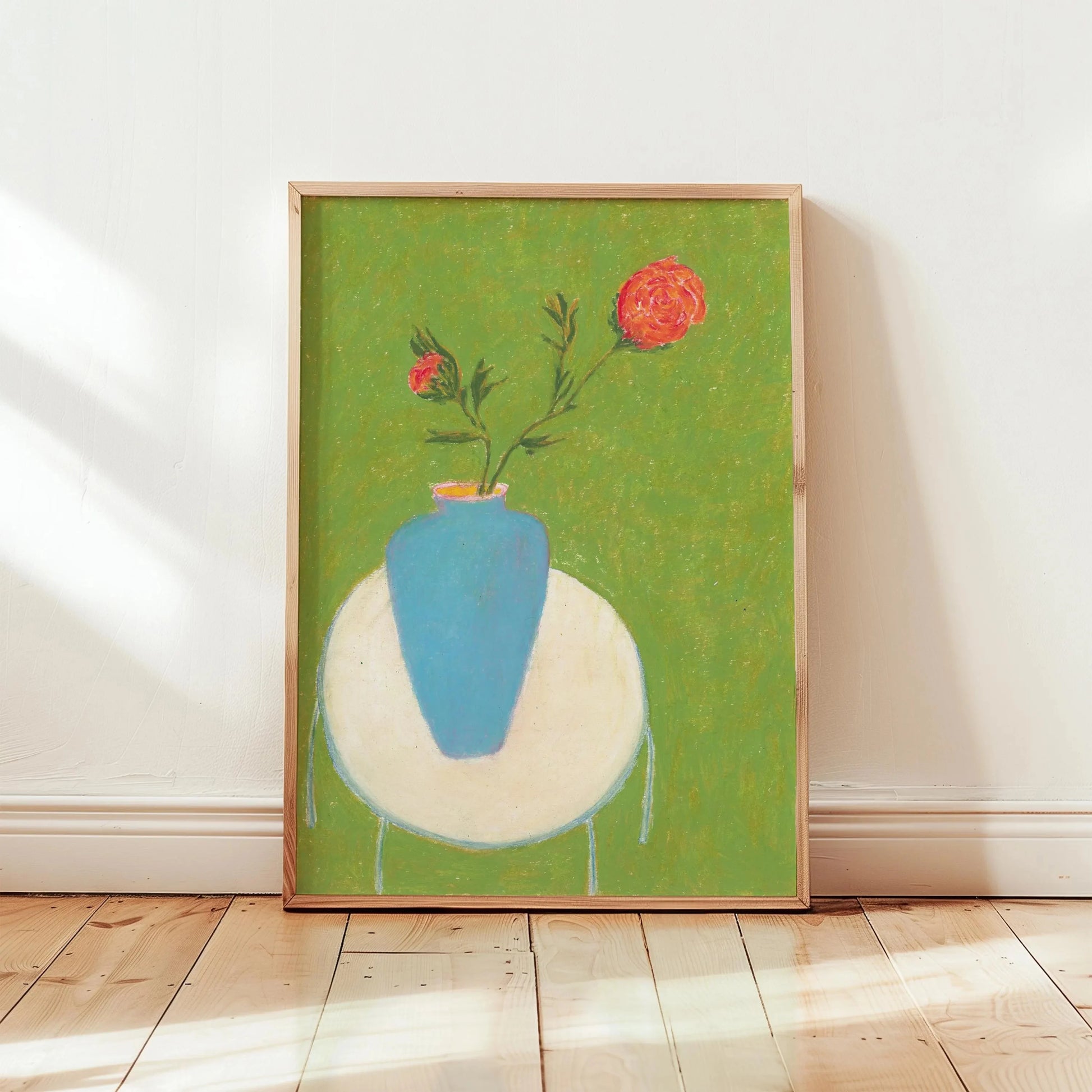 Framed artwork of a blue vase with red flowers on a green background, displayed on a wooden floor.