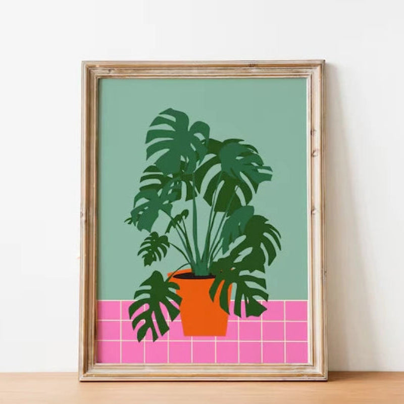 Framed artwork of a potted plant on a green background with pink tiles, displayed on a wooden surface.