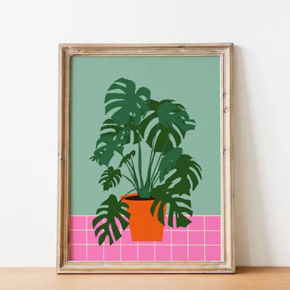Framed artwork of a potted plant on a green background with pink tiles, displayed on a wooden surface.