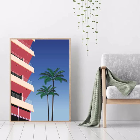 Framed artwork of a building and palm trees in a room with a chair and plant. Canvas & Curio