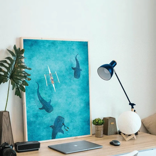 Desk with a laptop, lamp, and decorative poster of sharks and kayaks on a blue background. Canvas & Curio