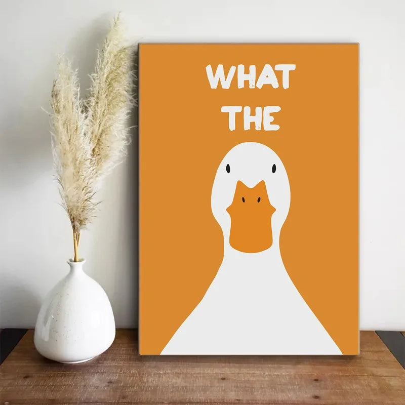 Canvas print with a duck illustration and 'WHAT THE' text on an orange background, placed on a wooden surface next to a white vase with dried plants.